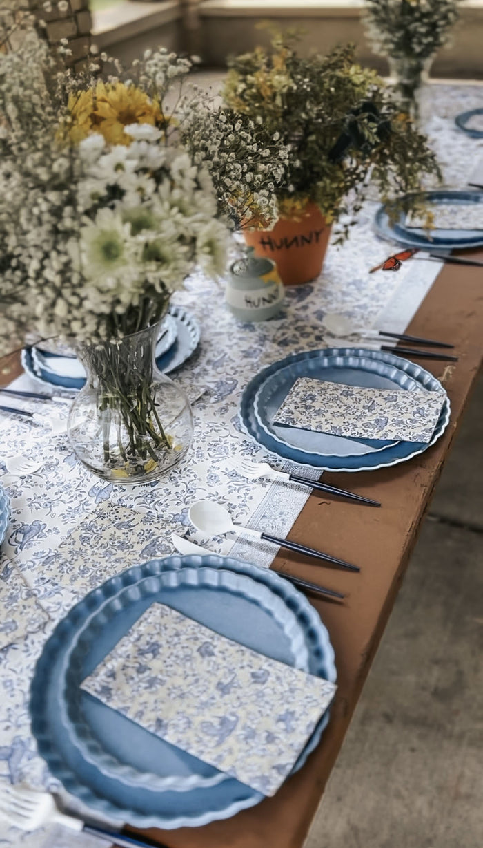 A table set for a meal features blue plates, Hester & Cook Blue Regal Peacock Guest Towel Napkins, dark cutlery, and a white and blue floral runner. Vases with white and yellow flowers add casual elegance to the gathering.