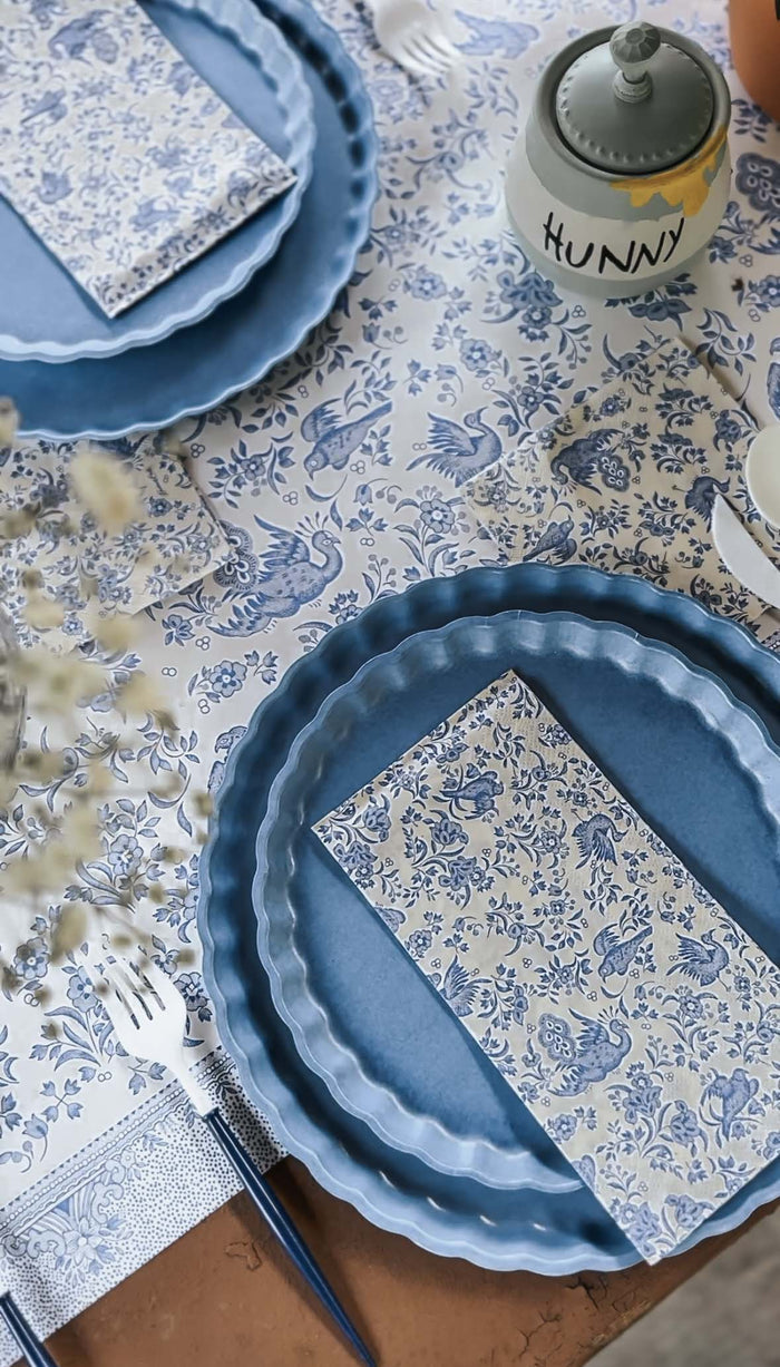 A table set with blue plates, forks, and Hester & Cook’s Blue Regal Peacock Guest Towel Napkins on a blue and white floral tablecloth, creating an elegant setting with coordinated details and a charming color scheme.