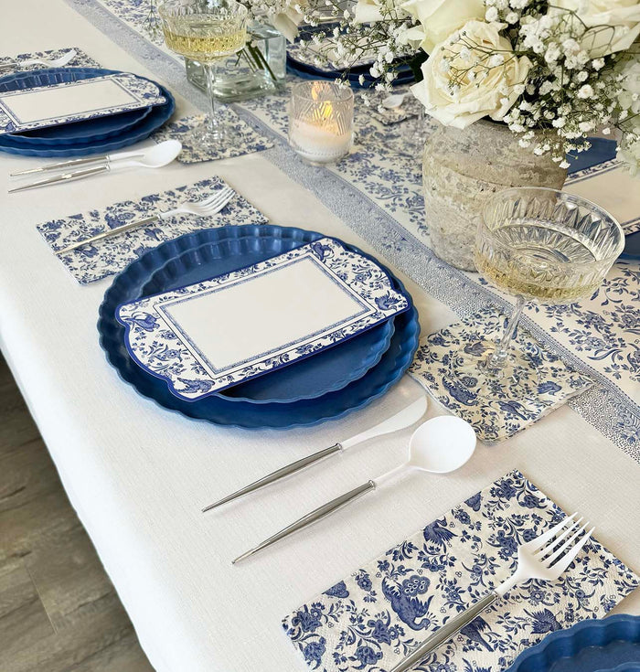 Elegant table setting with blue and white floral plates, Hester & Cook Blue Regal Peacock Guest Towel Napkins (16pk), place cards, crystal glasses, white cutlery, candles, and a vase of white flowers on a crisp white tablecloth.