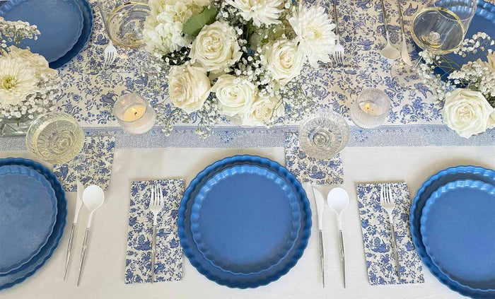 A table set for a meal features blue plates, white plastic cutlery, and Hester & Cook Blue Regal Peacock guest towel napkins. The centerpiece is white roses with small candles on a blue floral Hester & Cook table runner.