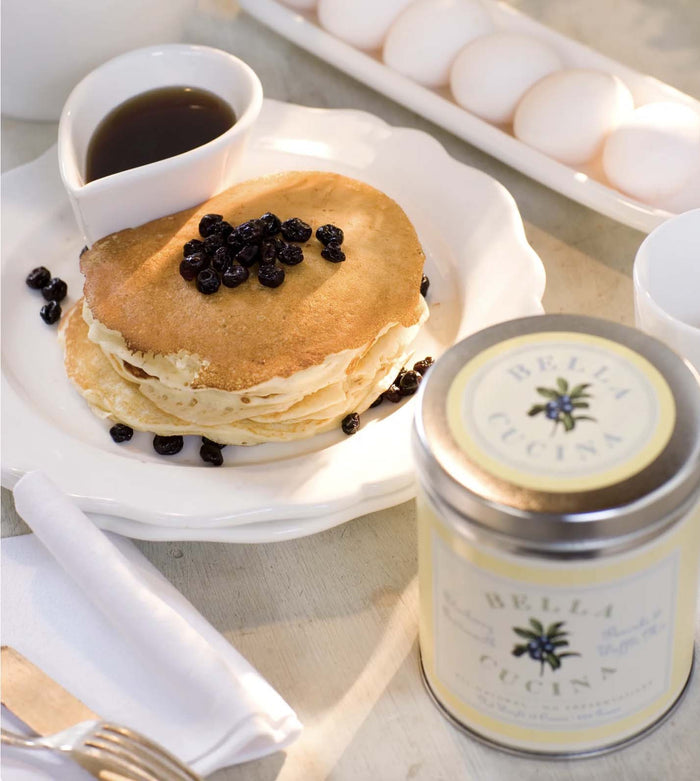 A stack of pancakes made with Bella Cucina’s Blueberry Buttermilk Pancake & Waffle Mix sits on a white plate topped with blueberries, next to a heart-shaped dish of syrup, fresh eggs, cutlery on a napkin, and a Bella Cucina canister.
