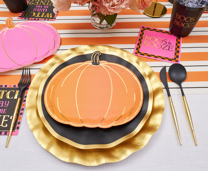 A festive table set with Sugarplum Boo Bold Assorted Die Cut Salad Plates, gold and black utensils, striped placemats, and “BOOS & BOOZE” Halloween napkins. Pink and orange flowers in a vase add a cheerful holiday accent.