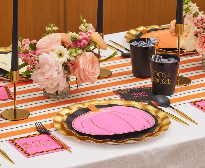 A festive table setting with Sugarplum’s Boo Bold Assorted Die Cut Salad Plates (8pk), pink pumpkins, a striped runner, black candles, floral arrangements in pink and white, and party cups labeled BOOS & BOOZE.
