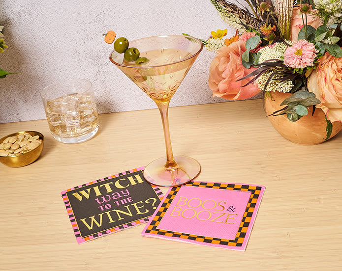 A martini with olives sits beside a whiskey, nuts, flowers, and Sugarplum Boo Bold Cocktail Napkins (26pk)—perfect accents for your Halloween party.