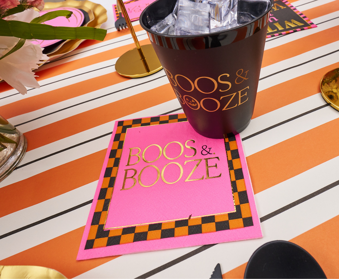A black cup with ice and Sugarplum's Boo Bold Cocktail Napkins (26pk) sit on an orange, white, and black striped table beside Halloween-themed decor.