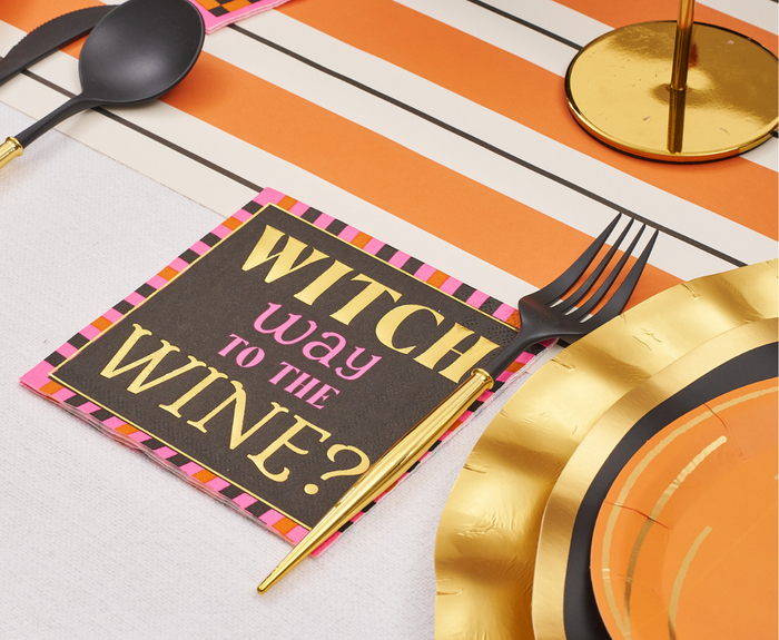 A Halloween party table features Sugarplum Boo Bold Cocktail Napkins (26pk) with "WITCH WAY TO THE WINE?" in gold and pink, paired with gold and black utensils, orange-striped decor, and gold-rimmed plates.
