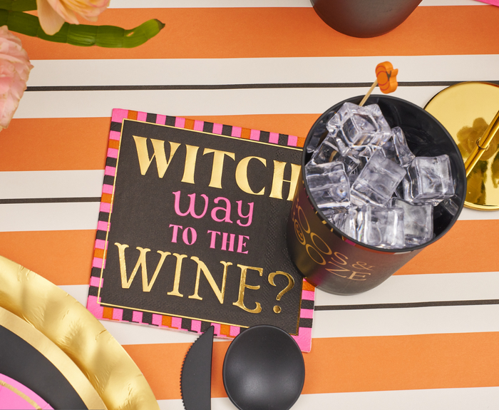 The Sugarplum Boo Bold Cocktail Napkins B/26pk, featuring "Witch way to the wine?" text, sit on a striped orange and white tablecloth with gold plates, ice cubes, and a pink flower—perfect for your next Halloween party.