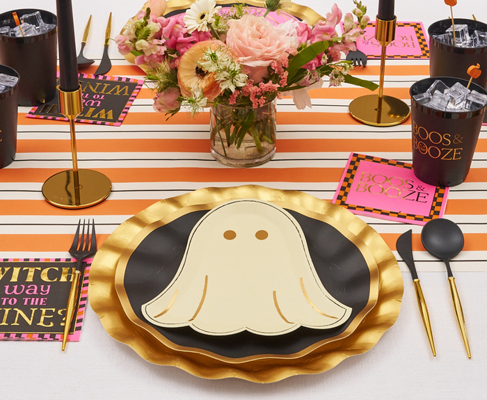 A festive Halloween table features Sugarplum Boo Bold Die Cut Salad Plates layered on gold foil plates, with black and gold cutlery, themed cups and napkins. A floral centerpiece plus a striped runner add colorful flair.