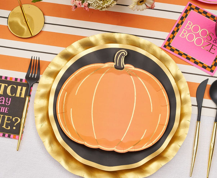 The Sugarplum Boo Bold Table Setting features a gold charger, black plate, pumpkin-shaped paper plate, black cutlery, and a "BOOS & BOOZE" napkin—all styled on a striped cloth for a festive Halloween look.