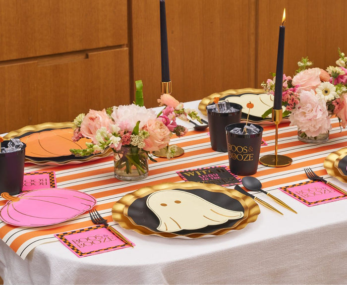 The Sugarplum Boo Bold Table Setting features a striped runner, pumpkin and ghost plates, gold-rimmed chargers, black BOOS & BOOZE cups, pink floral centerpieces, and tall black candles for a chic Halloween vibe.