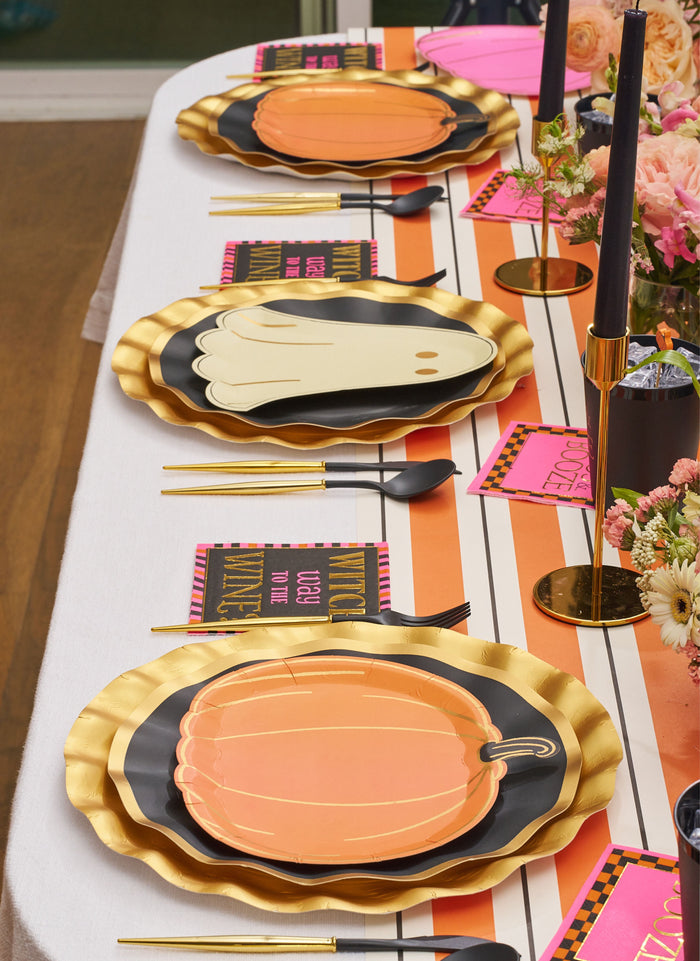 The Boo Bold Table Setting by Sugarplum showcases designer ghost and pumpkin plates, gold-rimmed accents, gold cutlery, black candles, pink napkins, and floral centerpieces on a striped white-and-orange tablecloth.