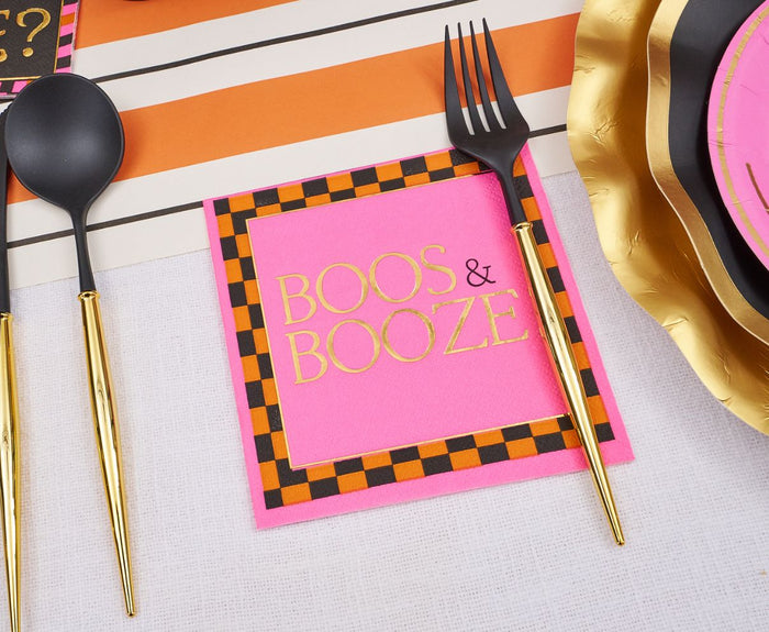 The Boo Bold Table Setting by Sugarplum features a pink napkin with a black and orange checkered border and gold "BOOS & BOOZE" lettering, plus designer plates and black utensils with gold handles on striped Halloween tableware.