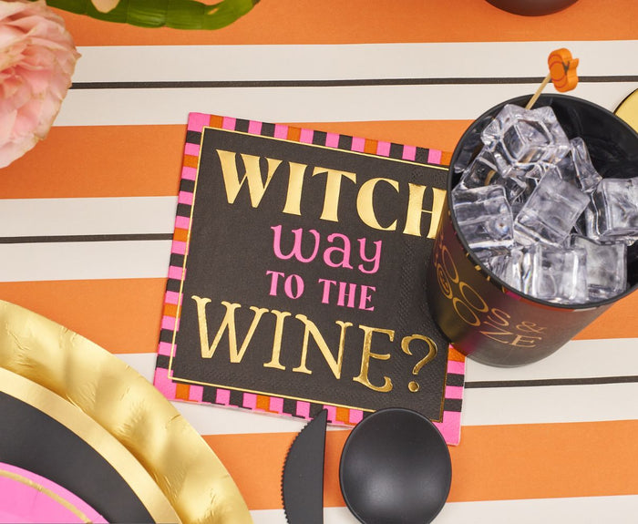 Create a festive Halloween display with the Sugarplum Boo Bold Table Setting, featuring a "Witch way to the wine?" napkin in gold and pink, a black cup with ice, designer plates, and an orange-striped tablecloth.