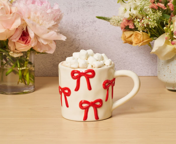 The Sophistiplate LLC Bow Mug + Hot Cocoa Gift Set is displayed on a wooden table between two vases of assorted flowers, creating a cozy and inviting scene.