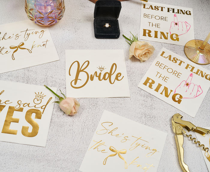 A flat lay of bridal shower items, including Sugarplum Bride Cocktail Napkins (26pk), gold-lettered cards (“Bride,” “She said YES,” “Last Fling Before the Ring”), a diamond ring in a box, two roses, a gold corkscrew, and a glass.