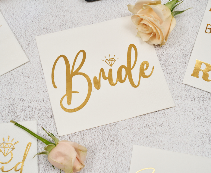 The Sugarplum Bride Cocktail Napkins (26pk) feature elegant gold script and a diamond icon, pairing perfectly with bridal shower décor. Shown surrounded by light peach roses on a textured surface.
