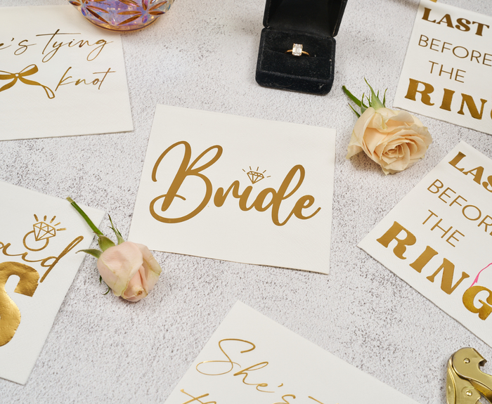 A flat lay displays Sugarplum Bride Cocktail Napkins (26pk), bachelorette-themed napkins, roses, and an engagement ring in a black box on a light surface.