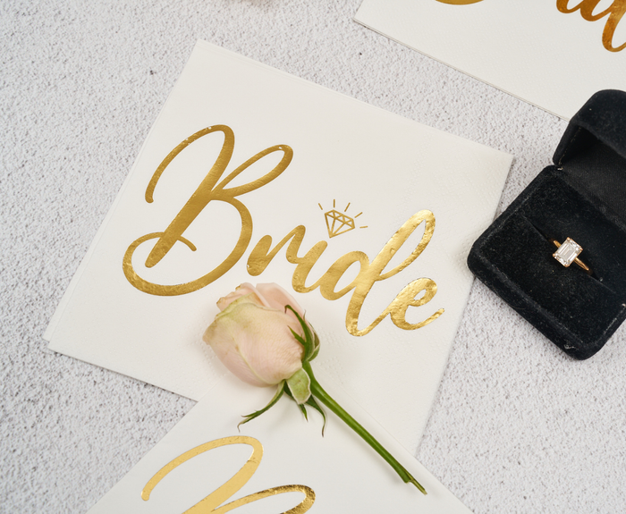 The Sugarplum Bride Cocktail Napkins (26pk) in white with gold script are styled with a diamond design, pale pink rose, and black ring box with a gold ring on a light textured surface.