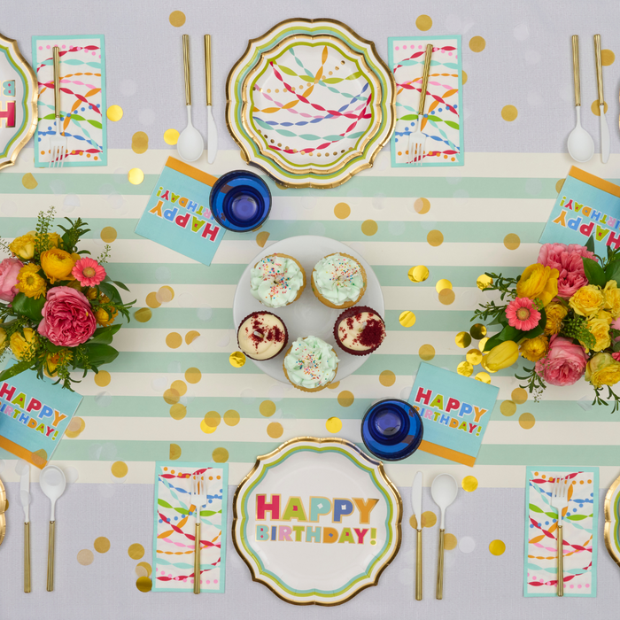 Create a stylish party with the Sophistiplate Bright Birthday Table Setting—designer disposable plates, colorful napkins, cupcakes, blue cups, gold confetti, and striped tablecloth for a festive tablescape.