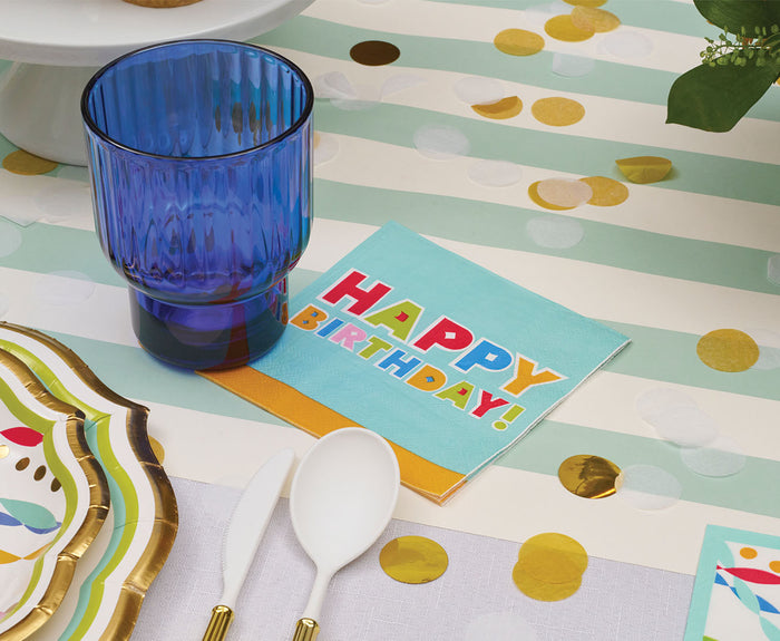 The Sophistiplate Bright Birthday Table Setting features a blue glass, "Happy Birthday!" napkin, white plastic spoons, designer plates with gold trim, and gold confetti on a striped tablecloth for a stunning disposable tablescape.