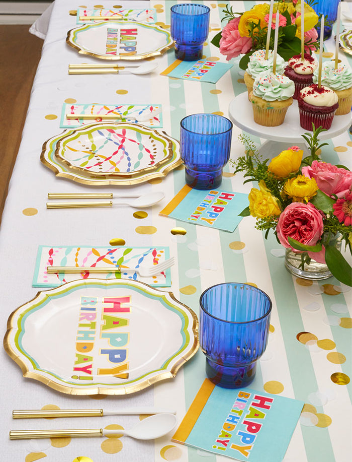 The Sophistiplate Bright Birthday Table Setting features designer disposable plates, blue cups, gold cutlery, cupcakes with candles, and a pink & yellow flower bouquet on a white tablecloth with gold polka dots.