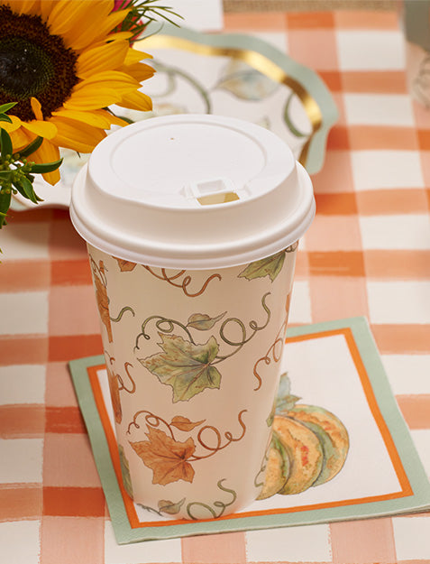 A disposable coffee cup with a white lid and autumn leaf design rests on Sophistiplate’s Bright & Thankful Cocktail Napkin (20pk) atop an orange and white checkered tablecloth, with a sunflower hinting at an autumn gathering.