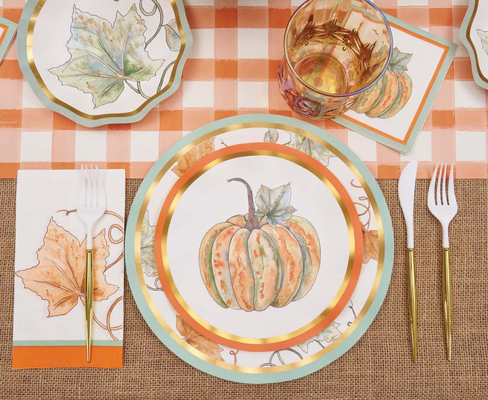 A table set for seasonal gatherings features a pumpkin-themed plate, Bright & Thankful Guest Towels by Sophistiplate, a leaf-patterned placemat, gold and white cutlery, a glass, and an orange-and-white gingham tablecloth.