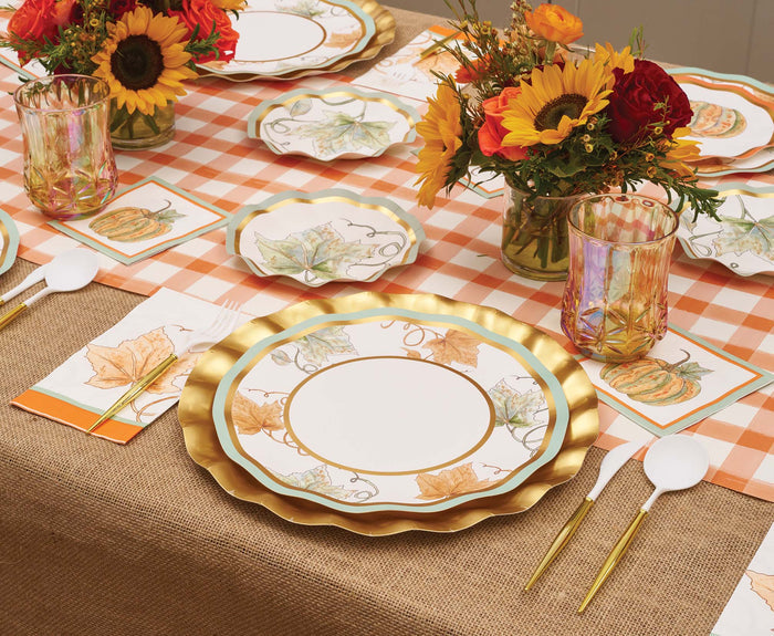 A festive autumn table setting featuring Sophistiplate’s Bright & Thankful Dinner Plates (8pk), each adorned with metallic gold trim, complemented by floral centerpieces, pumpkin-themed disposable plates, elegant gold cutlery, and an orange plaid table runner atop a burlap tablecloth.