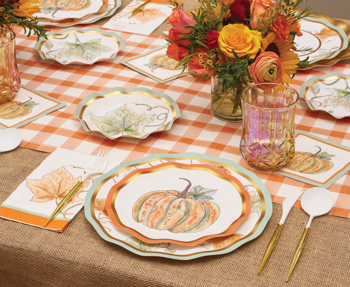 A festive autumn table is set with Sophistiplate’s Bright & Thankful Salad Plates (8pk), featuring pumpkin and leaf-themed gold trim, complemented by gold utensils, an orange plaid tablecloth, and glasses. The centerpiece showcases floral arrangements of orange and yellow blooms.