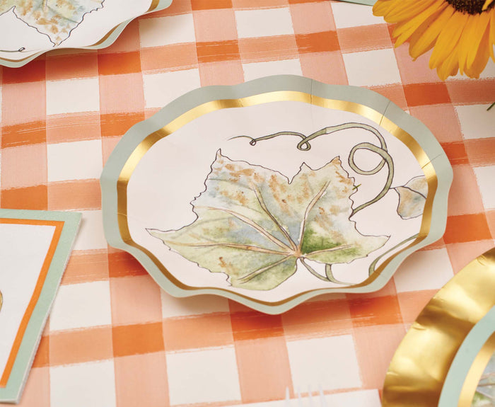 The Bright & Thankful Table Setting by Sophistiplate features a scalloped disposable plate with a gold rim and watercolor leaf design, set on a peach and white checkered tablecloth for an inviting autumn look with sunflower accents.
