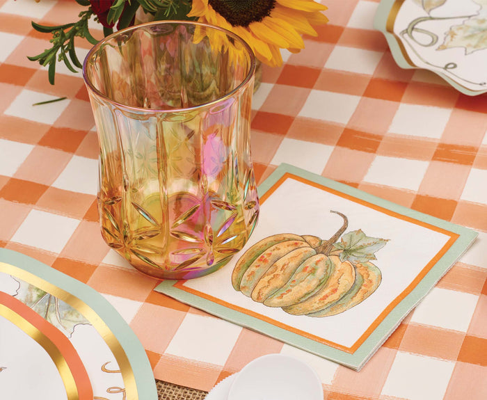 The Bright & Thankful Table Setting by Sophistiplate features a faceted amber glass on a watercolor pumpkin napkin, atop an orange and white checkered tablecloth, with designer plates and festive sunflower-themed tableware for autumn charm.