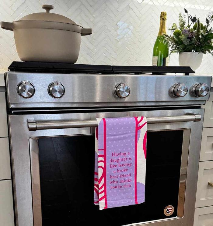 The Broke Best Friend Dish Towel by Drinks On Me hangs on a stainless steel oven with a pot on the stove, displaying the funny message: “Having a daughter is like having a broke best friend who thinks you’re rich.”.