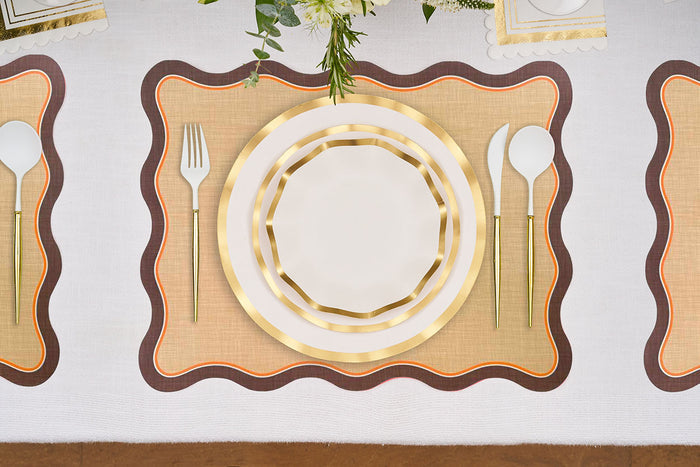 A table setting features Rosanne Beck’s Brown & Orange Scallop Edge Placemats (10pk) with gold-accented plates and gold-handled flatware, all atop a white tablecloth and topped with a floral centerpiece.