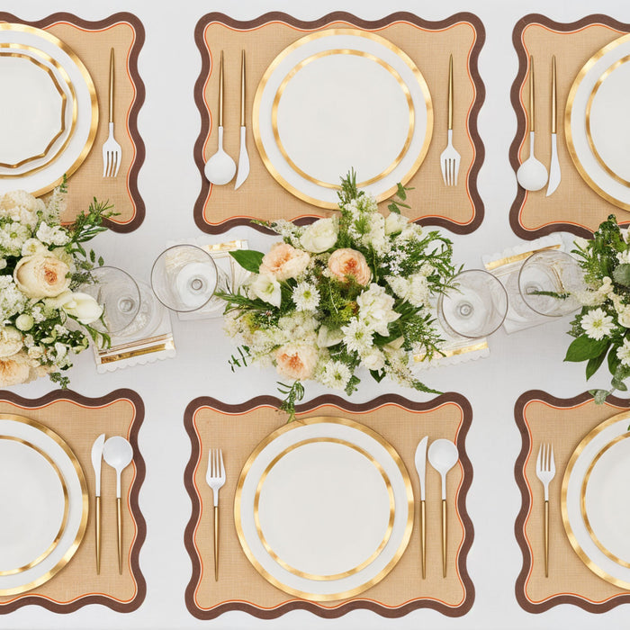 A top-down view of an elegant table setting featuring Rosanne Beck Brown & Orange Scallop Edge Placemats (10pk), gold-rimmed plates, gold flatware, and white floral centerpieces on a white tablecloth.
