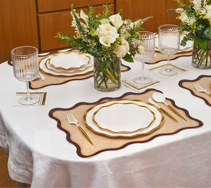 A dining table set for four showcases Rosanne Beck Brown & Orange Scallop Edge Placemats, white and gold-rimmed plates, gold cutlery, crystal glasses, and floral centerpieces in glass vases atop a crisp white tablecloth.