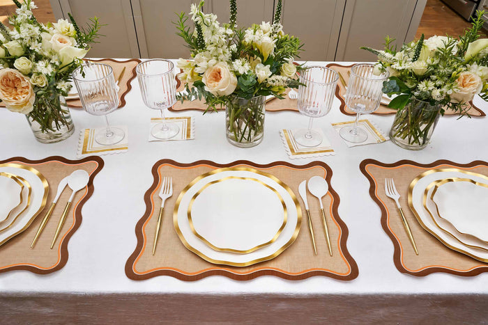 A stylish table features gold-rimmed plates, gold utensils, striped glassware, and white floral centerpieces atop a white cloth accented with Rosanne Beck Brown & Orange Scallop Edge Placemats (10pk) for an elegant touch.