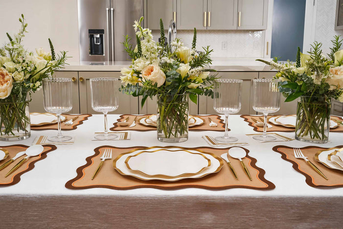 A modern dining table set for six with gold-rimmed plates, gold cutlery, Rosanne Beck Brown & Orange Scallop Edge Placemats, clear glasses, and three vases of white and peach flowers. Stainless steel kitchen appliances are visible in the background.