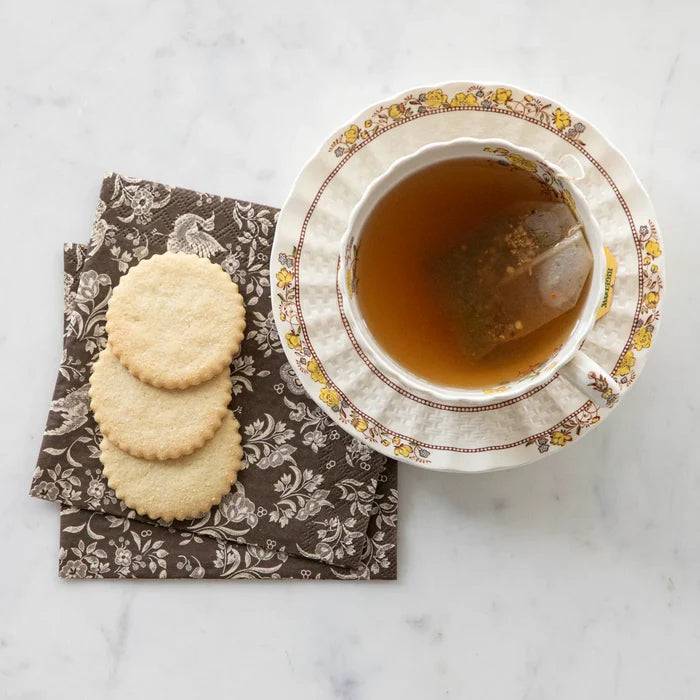 A teacup with tea and a teabag sits on a matching saucer next to three round cookies atop Hester & Cook Brown Regal Peacock Cocktail Napkins (20pk), all arranged on a white marble surface.