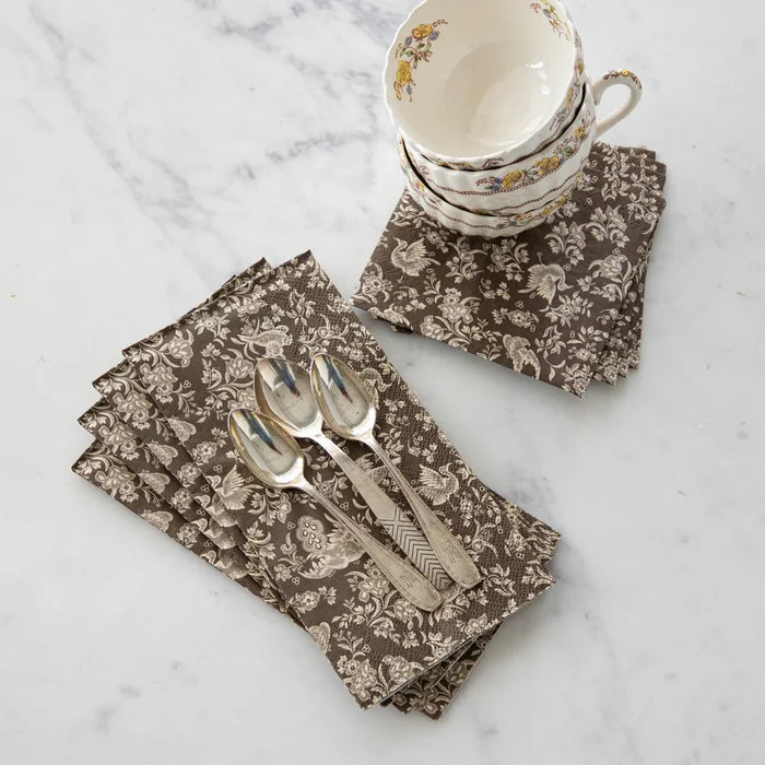 Three silver spoons rest on a stack of Hester & Cook Brown Regal Peacock Guest Napkins (16pk), with three floral teacups and saucers stacked nearby on a white marble surface.