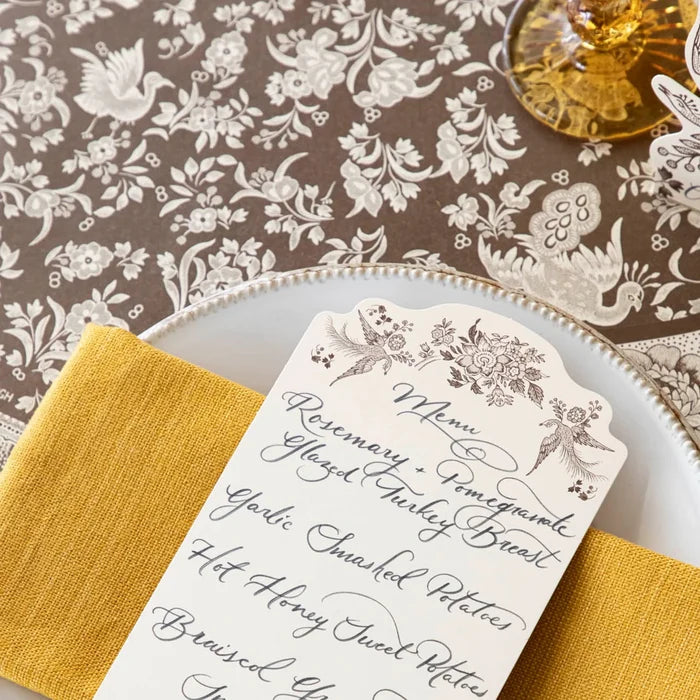A decorative menu card with elegant cursive and floral accents rests on a white plate with a yellow napkin, atop the Hester & Cook Brown Regal Peacock Runner (20x25").