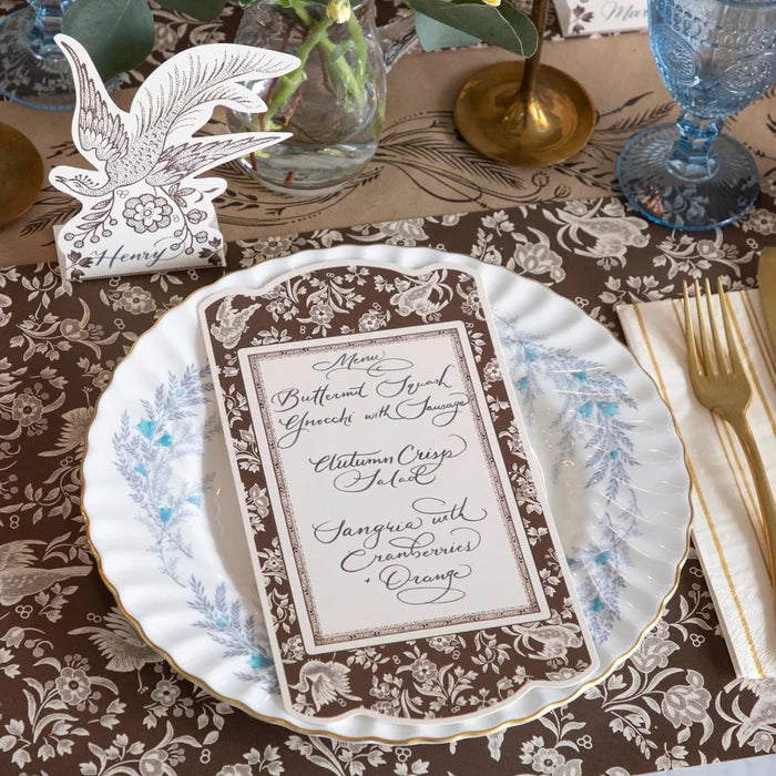 A close-up of an elegant table setting featuring a floral menu on a blue-and-white plate, gold cutlery, blue glassware, and a Brown Regal Peacock Table Card by Hester & Cook (12pk), all accented with autumn-themed décor.