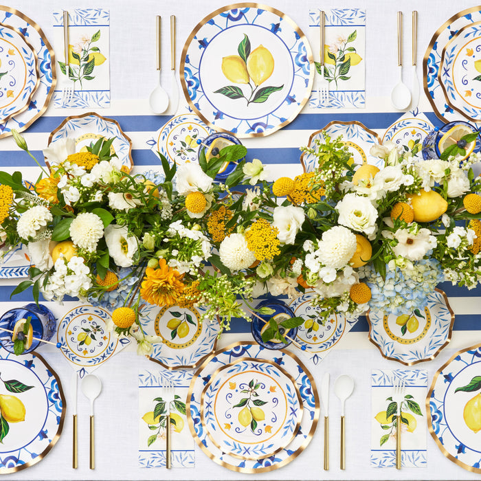 Create a festive table with the Capri Coast Table Setting by Sophistiplate LLC—heavy duty disposable party plates featuring lemon-inspired blue and white patterns, accompanied by white utensils and napkins neatly arranged for effortless and stylish entertaining. A centerpiece of yellow and white flowers completes the vibrant look.