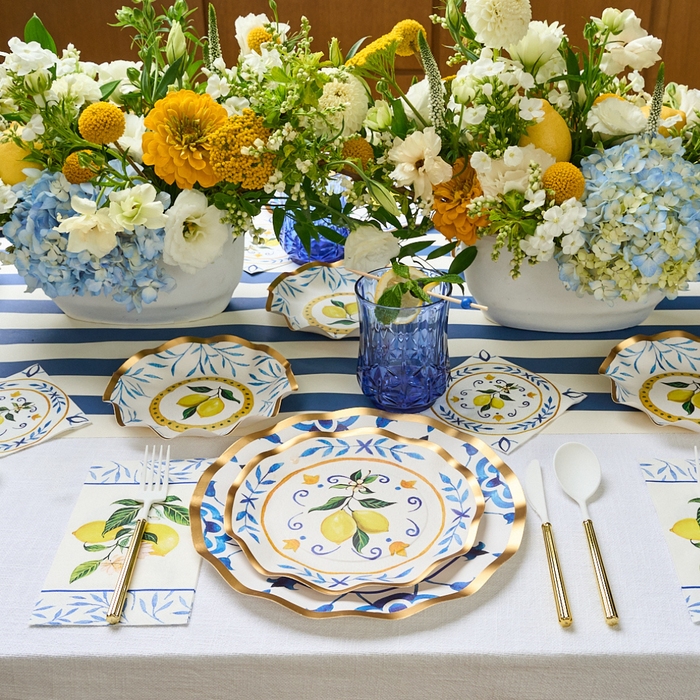 The Capri Coast Table Setting by Sophistiplate LLC features lemon-themed disposable plates, napkins, blue glasses, and gold utensils on a blue-and-white striped tablecloth, accented with yellow, white, and blue flower arrangements.
