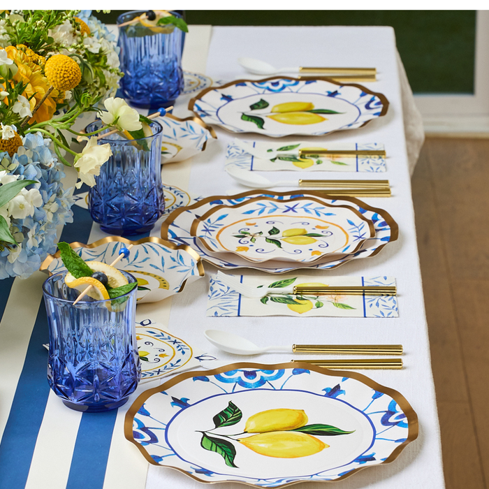 The Capri Coast Table Setting by Sophistiplate LLC features lemon-themed heavy duty paper plates, gold cutlery, blue glasses, a striped runner, and a white-yellow-blue floral centerpiece for an elegant, vibrant look.