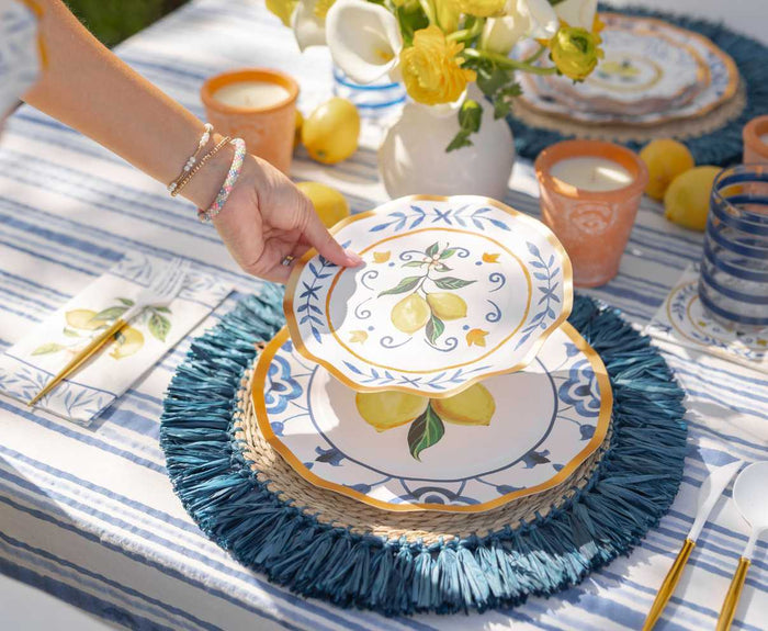 A hand places the Capri Coast Table Setting by Sophistiplate LLC—a lemon-themed heavy duty paper plate—on a blue placemat atop a picnic table decorated with blue stripes, yellow flowers, lemons, and matching tableware.