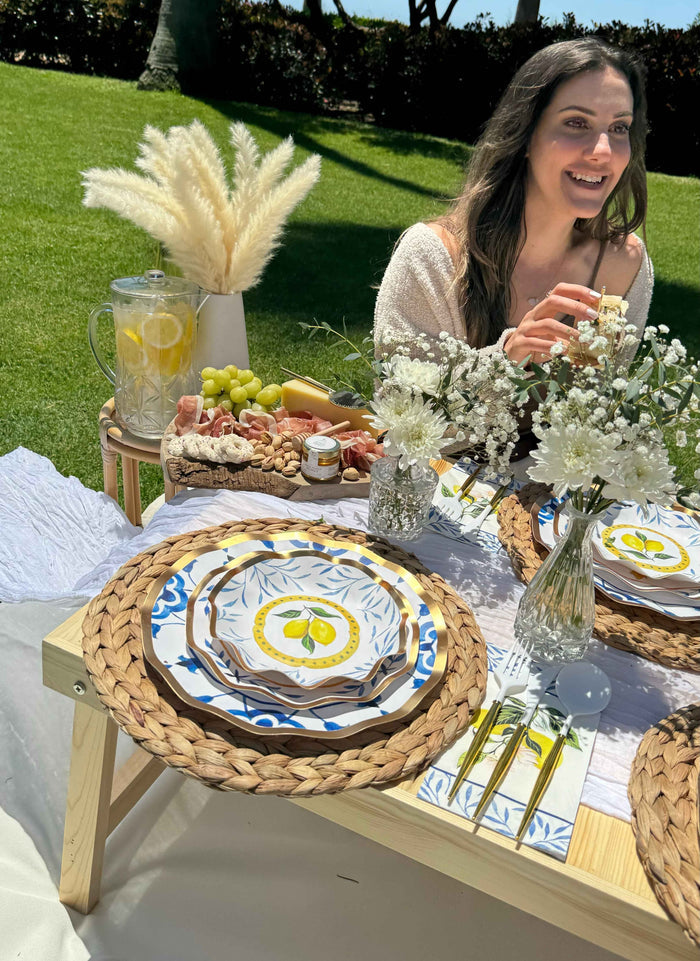 A woman smiles at an outdoor picnic table set with Sophistiplate LLC’s Capri Coast Table Setting—floral plates, woven placemats, white flowers in vases, snacks, and lemon water on a sunny grassy lawn.