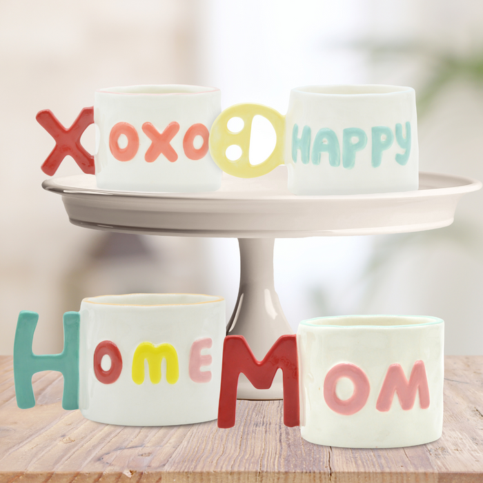 Four 14oz Ceramic Message Mugs by Sophistiplate, each with raised pastel letters spelling “XOXO,” “HAPPY,” “HOME,” and “MOM,” are artfully stacked on a white cake stand against a soft, blurred background.