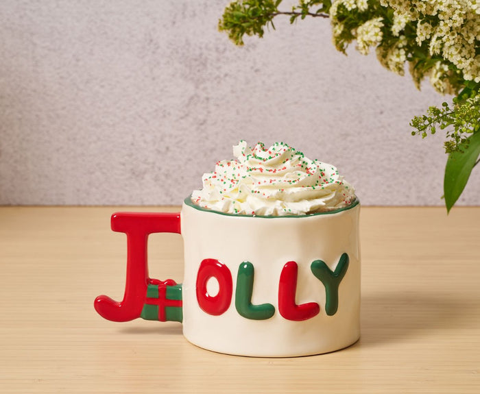 The Sophistiplate Ceramic Message Mug Jolly, 14oz, features colorful "JOLLY" lettering and a large red J handle. Brimming with whipped cream and sprinkles, it rests on a wooden surface beside a bouquet of flowers.