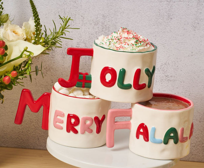 Three Sophistiplate 14oz Ceramic Message Mugs—each with festive words (“JOLLY,” “MERRY,” and “FALALA”)—form a pyramid and feature holiday toppings; flowers are seen to the left.