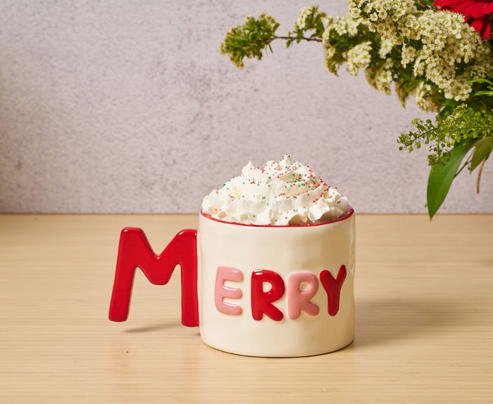 The Sophistiplate Ceramic Message Mug Merry, 14oz, features the word "MERRY" and a red M-shaped handle. Microwave and dishwasher safe. A floral arrangement with white flowers sits to the right.